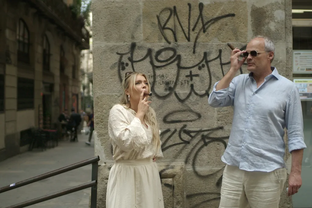 Rheborg och en kvinna står utomhus vid en stenmur täckt av graffiti och röker cigarr. Kvinnan bär en vit klänning och mannen är klädd i ljusblå skjorta och shorts. En stadsgata syns i bakgrunden.