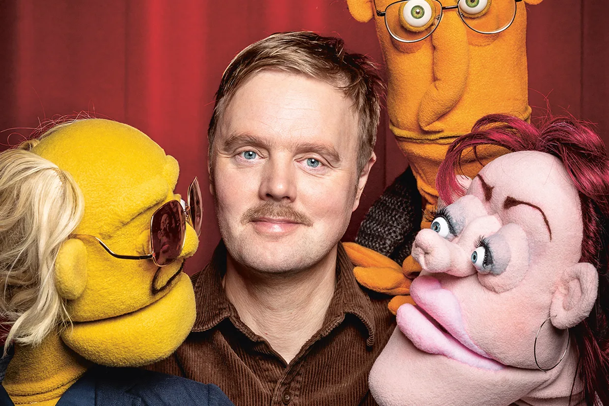 Björn Carlberg med kort blont hår och mustasch står framför en röd gardin, omgiven av tre färgglada dockor med uttrycksfulla ansikten och olika frisyrer.