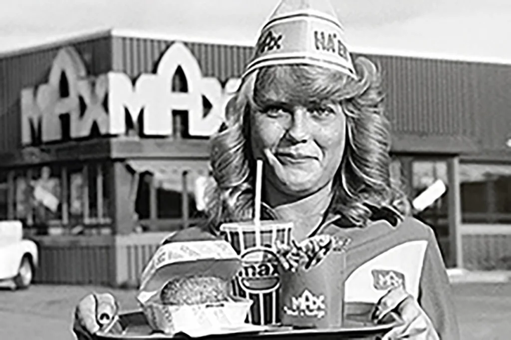 En kvinna i uniform och pappershatt håller en matbricka med en hamburgare, pommes frites och en drink framför en Max-restaurang. Bilden är svartvit och har en retrokänsla.