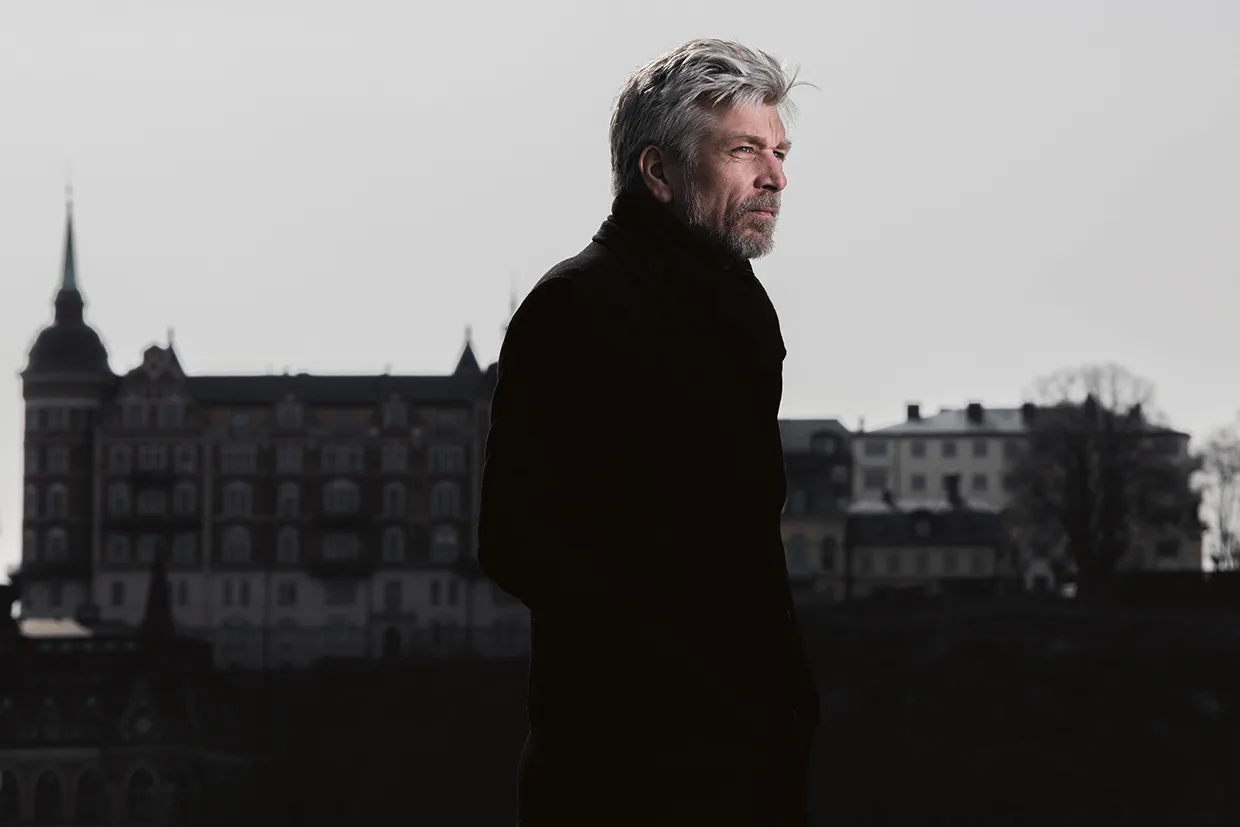 En man med grått hår och skägg står utomhus i profil, klädd i en mörk rock. Bakom honom syns en stor, historisk byggnad med flera fönster och en spetsig spira mot en grå himmel.