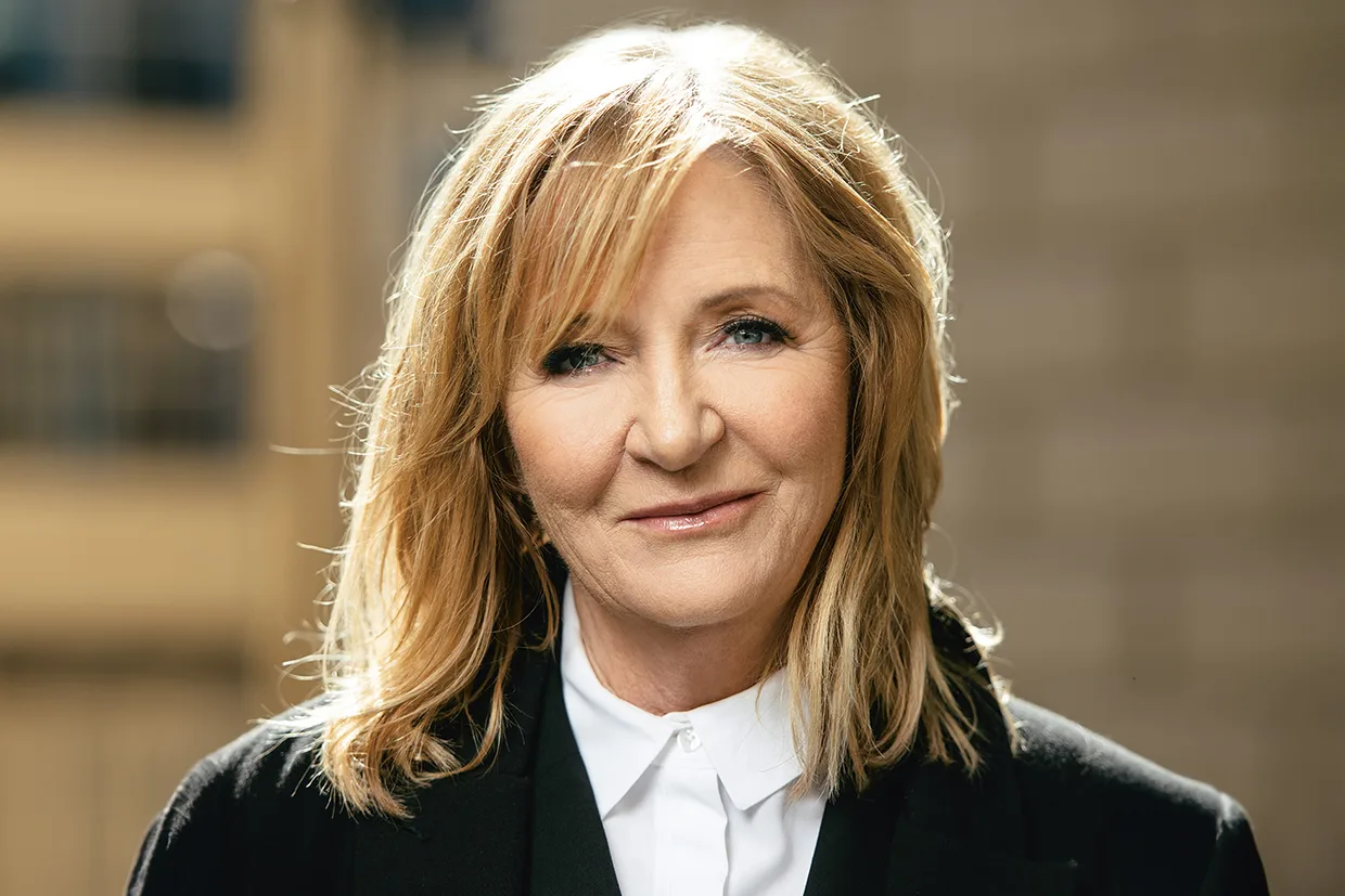 Malou Von Sivers med axellångt blont hår, klädd i en svart kavaj över en vit skjorta med krage, ler mjukt medan hon står utomhus i naturligt ljus med en suddig bakgrund.