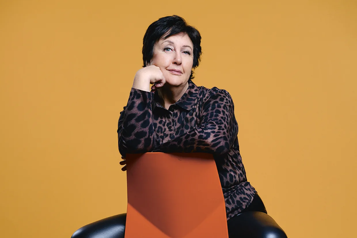 Babben Larsson med kort mörkt hår, klädd i leopardmönstrad blus och svarta byxor, sitter bakåtlutad på en orange stol och vilar hakan på handen mot en enfärgad gul bakgrund.