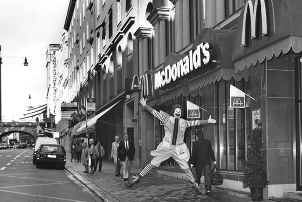 En person utklädd till Ronald McDonald hoppar med armar och ben utspridda utanför en McDonalds-restaurang på en gata i en stad, medan fotgängare går förbi. Bilden är i svartvitt.
