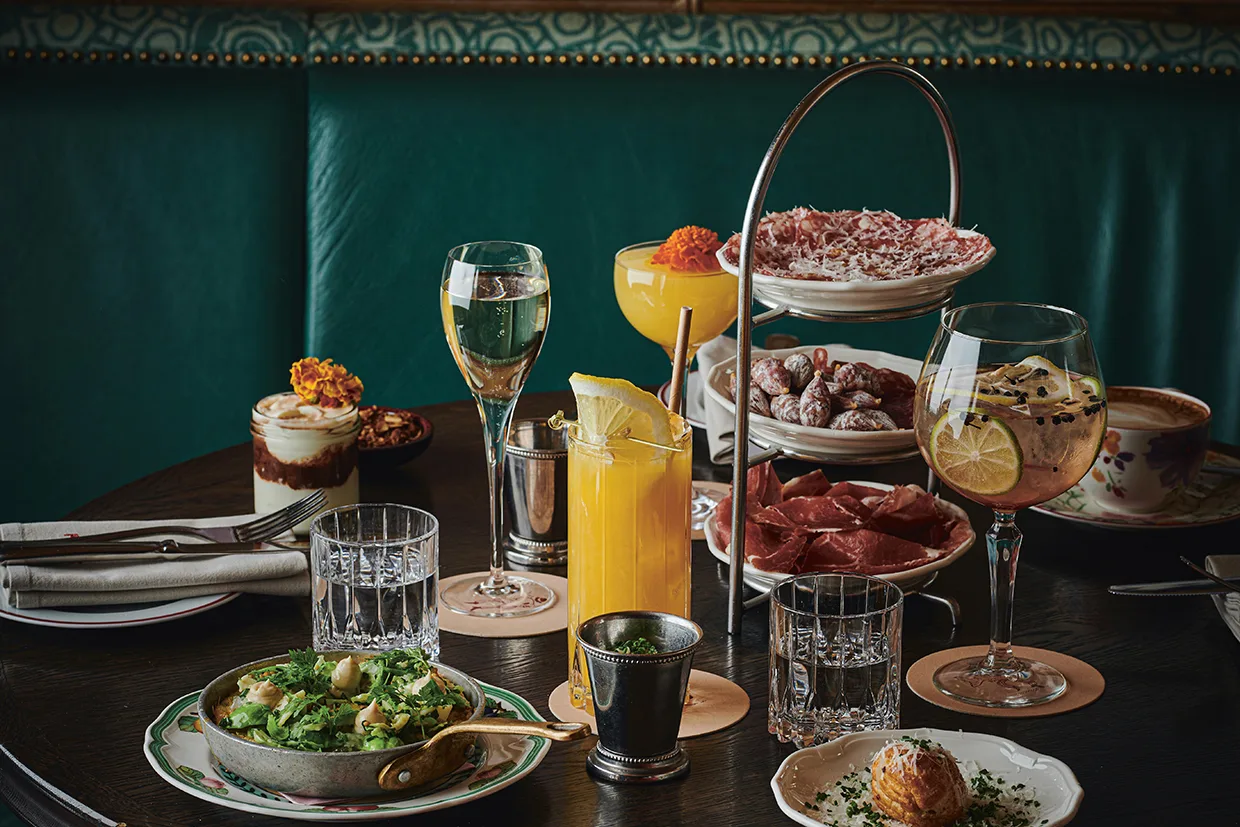 Ett bord dukat med cocktails, vin, vatten och juice, omgivet av olika gourmeträtter som charkuterier, bröd, sallad, dessert och små aptitretare mot en grön fond.