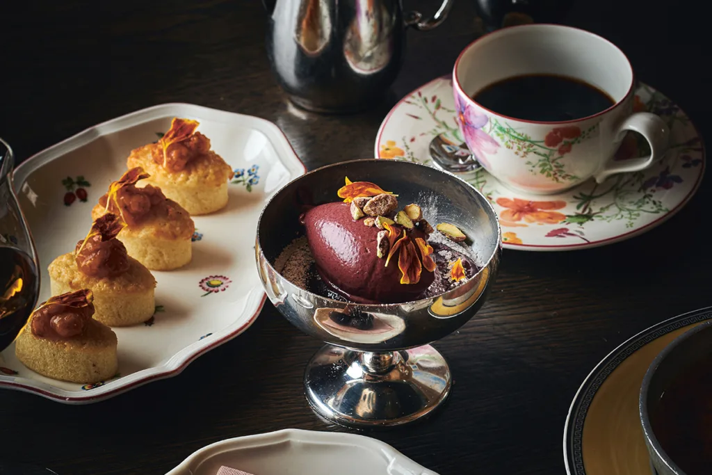 Ett silverfat med en skopa mörk bärsorbet garnerad med nötter och kronblad, bredvid en tallrik med små kakor toppade med karamell och krispiga skärvor, och en blommig tekopp fylld med svart kaffe.