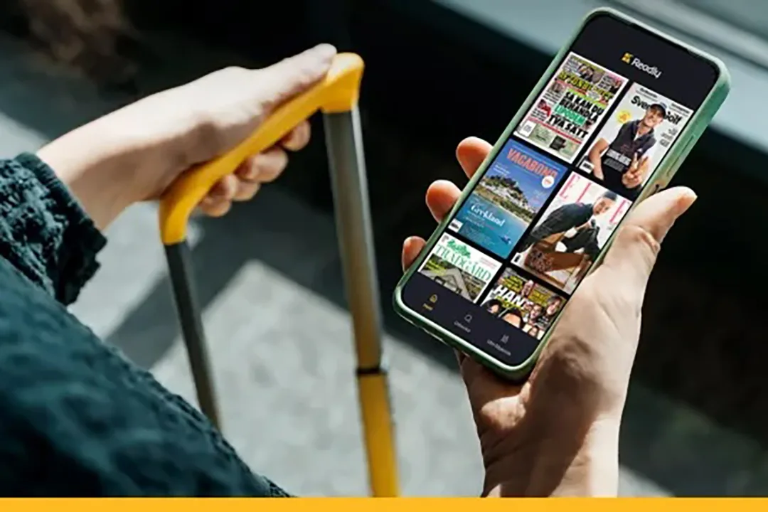 En person håller ett gult resväskhandtag i ena handen och en smartphone i den andra och visar en streaming- eller biblioteksapp med olika tidningar och publikationer på skärmen.
