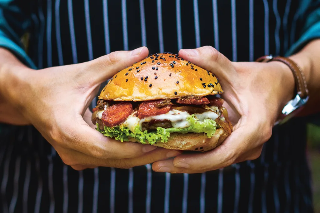 En person i randigt förkläde håller en stor hamburgare med sallad, smält ost, skivat kött och ett sesamfröbröd i båda händerna.