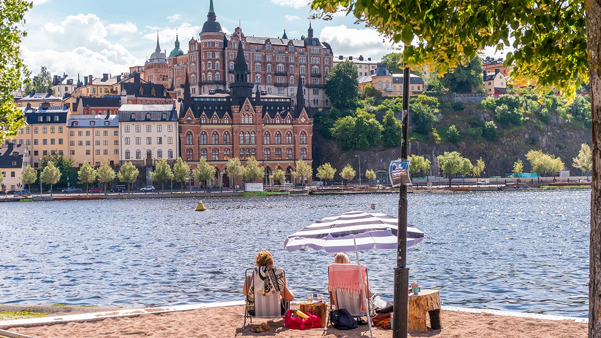 Två personer sitter under ett randigt parasoll på en sandstrand vid vattnet, med utsmyckade historiska byggnader och grönska som syns över vattnet under en delvis molnig himmel.
