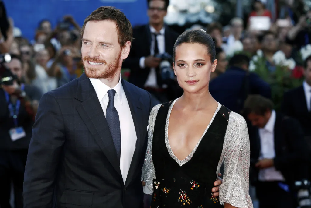 Michael Fassbender i mörk kostym och slips står bredvid Alicia Vikander i en djupt V-ringad klänning med silverfärgade ärmar på ett formellt utomhusevenemang, omgiven av en mängd människor och fotografer.