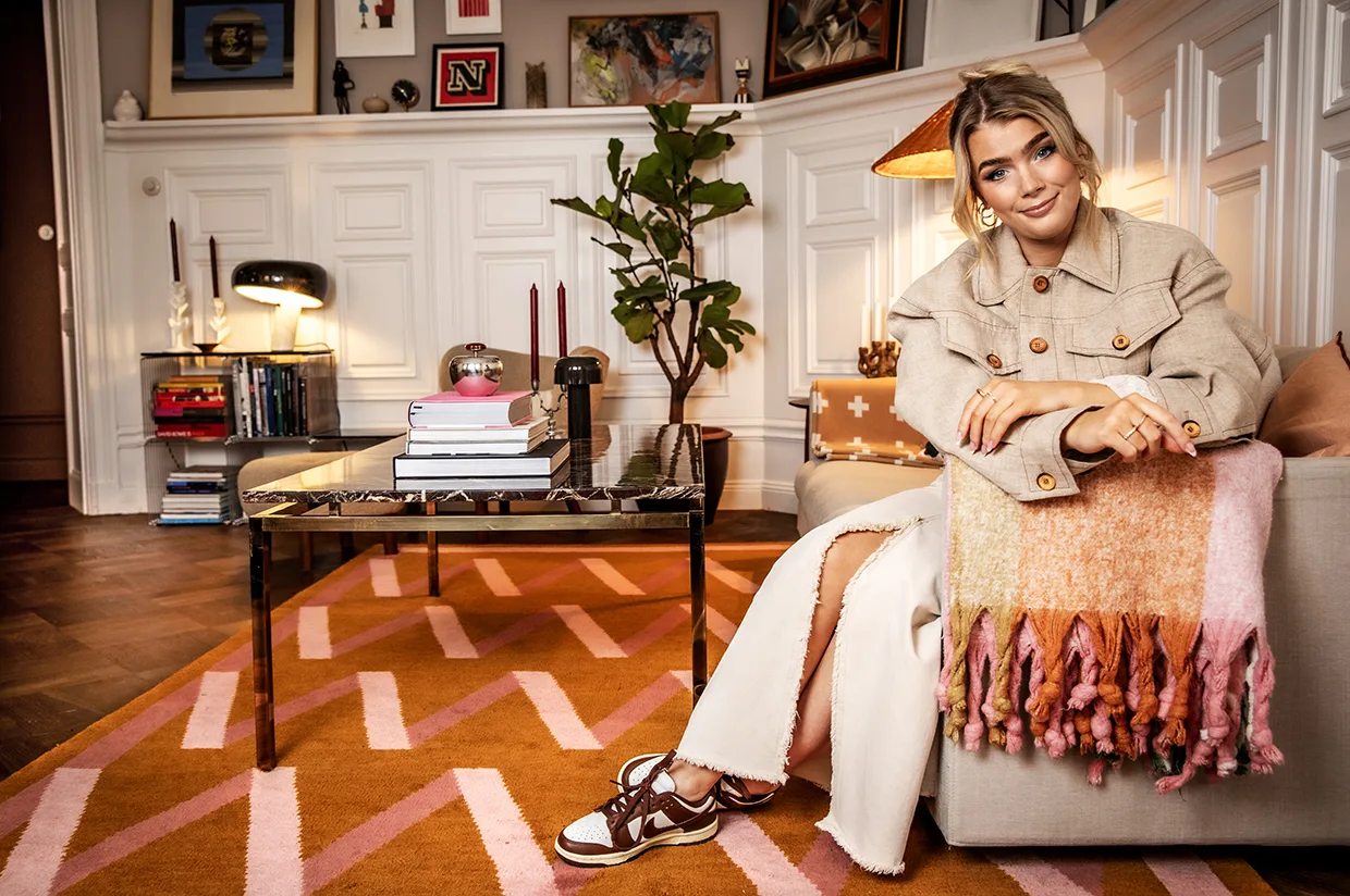 Keyyo sitter i en beige soffa med en färgglad filt, klädd i en brun kappa och sneakers. Det eleganta vardagsrummet har en mönstrad matta, soffbord med böcker, växter och eklektisk väggkonst och inredning.