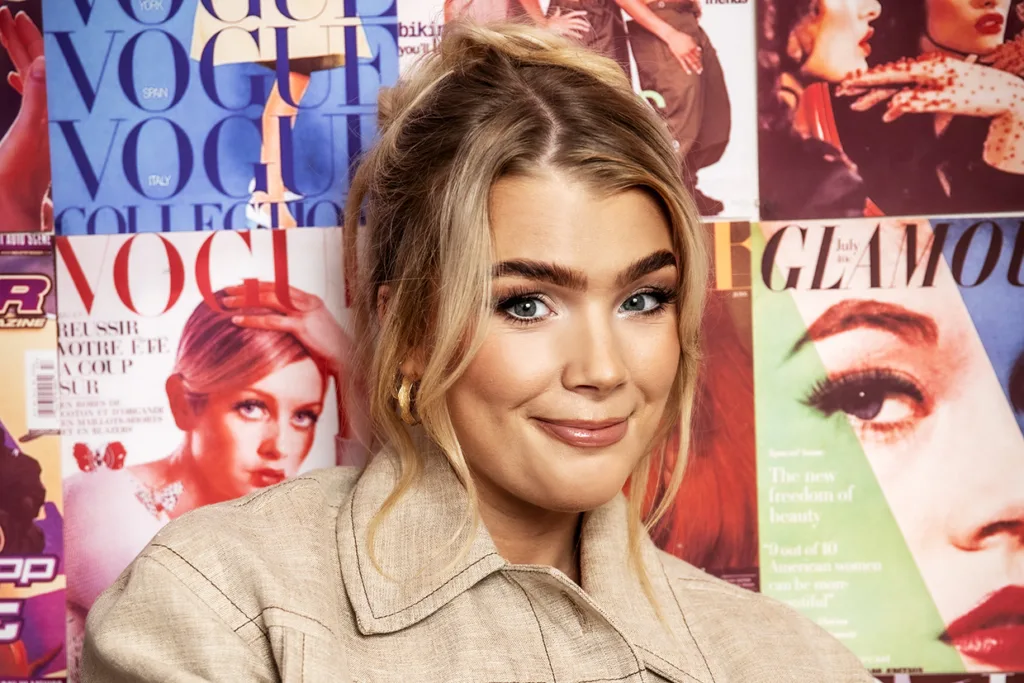 Keyyo med blont hår i en lös frisyr, klädd i en beige jacka, ler framför en färgglad vägg täckt av tidningsomslag från Vogue och Glamour.