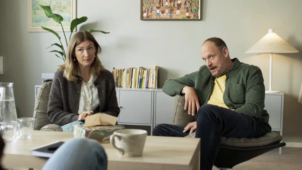 Sissela Benn och Jonatan Unge sitter i ett mysigt, väl upplyst vardagsrum, vända mot någon utanför kameran. Böcker, konst och en växt dekorerar bakgrunden. En kaffemugg och en vattenkanna står på bordet i förgrunden.