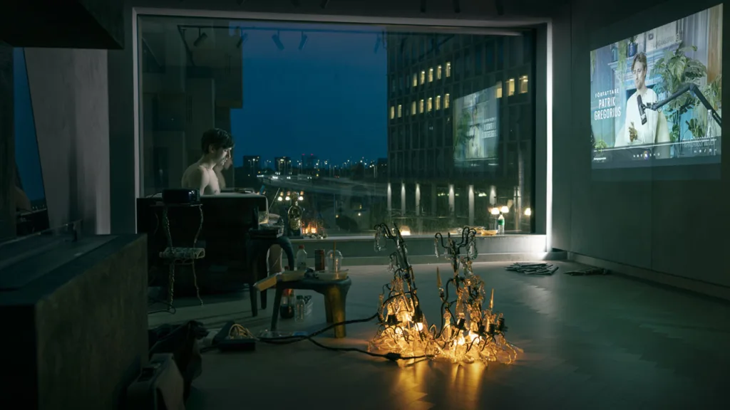 Isac Calmroth sitter vid ett skrivbord i en mörk lägenhet på kvällen, upplyst av mjuka lampor på golvet. Stora fönster avslöjar stadsbyggnader och gatlyktor utanför. En projicerad bild visas på väggen.