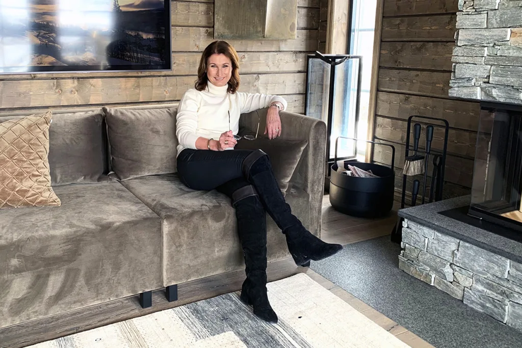 Viveca Sten sitter i en taupefärgad soffa i ett mysigt, modernt rum med träväggar och en öppen spis i sten. Hon bär en vit polotröja, svarta byxor och svarta knähöga stövlar och ler med ena armen vilande på soffans ryggstöd.
