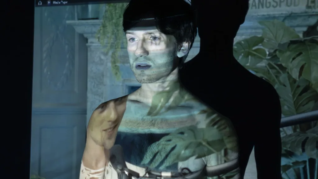 Isac Calmroth står framför en vägg med en projicerad bild av grönska och ett ansikte som överlappar hans kropp och ansikte, vilket skapar en visuell effekt med ljus och skugga.