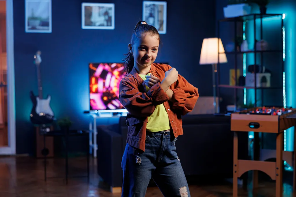 En ung flicka i brun jacka och slitna jeans poserar lekfullt med korsade armar i ett mysigt, neonbelyst rum med fotbollsspel, TV och gitarr i bakgrunden.