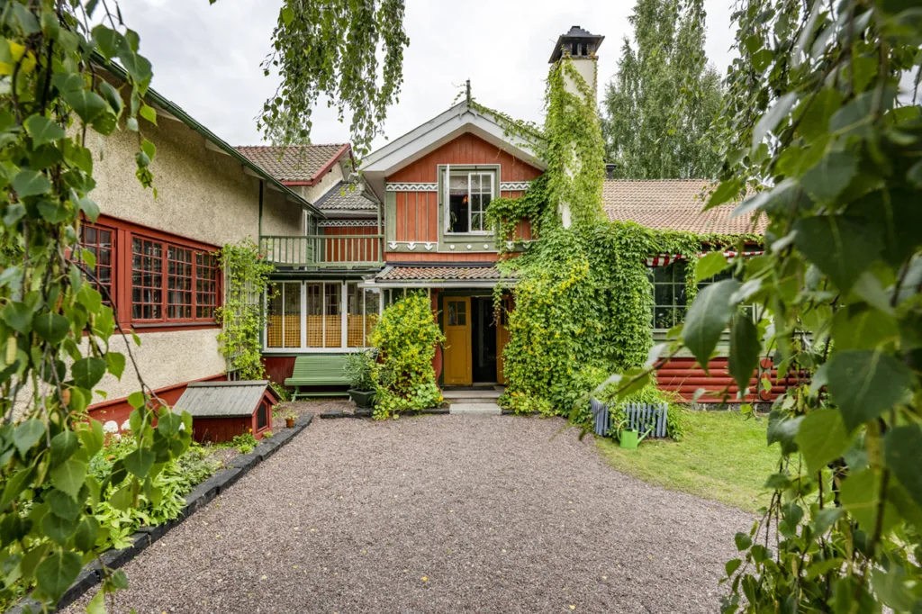 Ett charmigt, färgglatt hus, Carl Larsson gården, täckt av grön murgröna med röda och gula trädetaljer, omgivet av träd och grönska och med en grusad innergård framför.