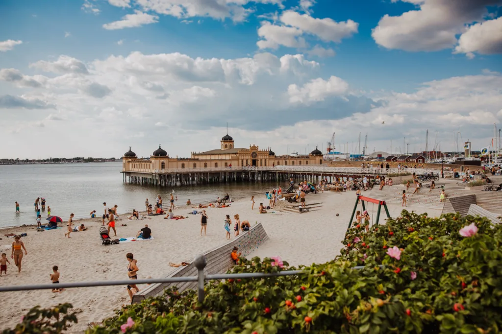 Människor kopplar av på en sandstrand nära en historisk pirbyggnad, Varbergkallbadhus, över vattnet. Himlen är delvis molnig och i förgrunden syns färgglada blommor och grönska.